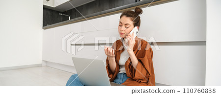 Woman looking bored and unamused while sitting on floor and listening to conversation, has laptop on laps, waiting on line, making phone call 110760838