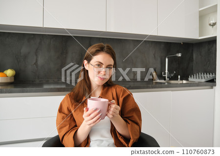 Portrait of happy young woman in glasses, sitting at home in kitchen, holding cup of coffee, drinking favourite tea, relaxing indoors on cold autumn day 110760873