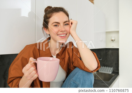 Beautiful young female model holding pink cup, sitting in kitchen and smiling at camera, woman drinking tea at home or enjoying aroma of coffee 110760875