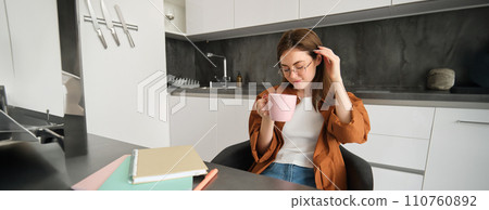 Portrait of young woman working from home, taking a break for cup of tea. Girl studies remotely, sits in kitchen, drinking hot coffee and smiles 110760892