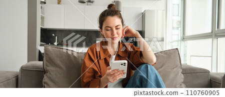 Portrait of happy young brunette woman sitting on sofa with smartphone, reading notification, messaging on mobile phone, resting on couch at home, smiling while looking at device screen 110760905