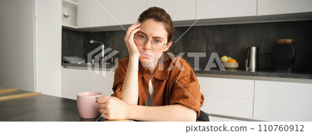 Portrait of young serious woman, working from home in glasses, sitting with folder and documents in kitchen, looking tired 110760912