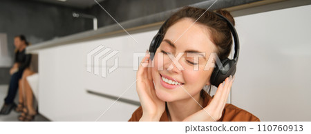 Image of modern woman, beautiful girl enjoys listening to music, has black wireless headphones on, sits on floor and rests Image of modern woman, beautiful girl enjoys listening to music, has black wireless headphones on, sits on floor and rests 110760913