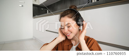 Cute brunette woman on kitchen floor, wears headphones, listens music and smiles, enjoys sound quality 110760935