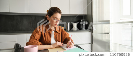 Portrait of woman looking concentrated, writing down notes, doing homework in kitchen, drinking coffee, studying at home 110760936