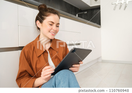 Portrait of pretty young woman at home, sitting on kitchen floor, looking at digital tablet, using gadget, reading on device 110760948