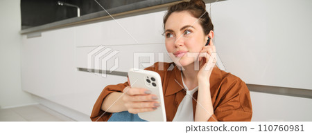 Portrait of young modern woman in wireless headphones, sitting on kitchen floor, using smartphone, making playlist with favourite songs, listens to music 110760981