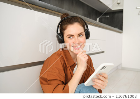 Positive young woman, sits at home with wireless headphones and smartphone, smiles and looks happy Positive young woman, sits at home with wireless headphones and smartphone, smiles and looks happy 110761008