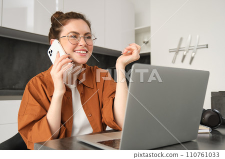 Portrait of young self-employed woman, entrepreneur working from home, freelancer calling client. Girl making an order, talking to someone on phone, sitting in kitchen with laptop 110761033