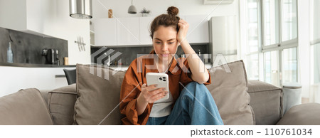 Portrait of young modern woman reading on mobile phone, scrolling social media app on smartphone, sitting on couch in living room in casual clothes Portrait of young modern woman reading on mobile phone, scrolling social media app on smartphone, sitting on couch in living room in casual clothes 110761034