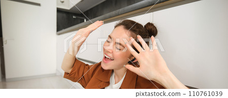 Carefree woman singing along song, listens to music in wireless headphones, enjoying sound quality, dancing with hands 110761039