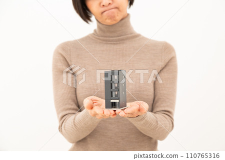 A middle-aged woman worries while holding a housing model of a building. 110765316