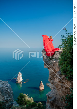 Red dress sea woman. Happy woman with flowing hair in a long flowing red dress stands on a rock near the sea. Travel concept, photo session at sea 110771946
