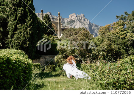 a beautiful woman with long brown hair and long white dress stay along a path along beautiful bushes in the park rear view a beautiful woman with long brown hair and long white dress stay along a path along beautiful bushes in the park rear view 110772096