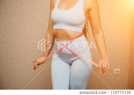 Cropped view of slim woman measuring waist with tape measure at home, close up. Unrecognizable european woman checking the result of diet for weight loss or liposuction indoors Cropped view of slim woman measuring waist with tape measure at home, close up. Unrecognizable european woman checking the result of diet for weight loss or liposuction indoors 110772136