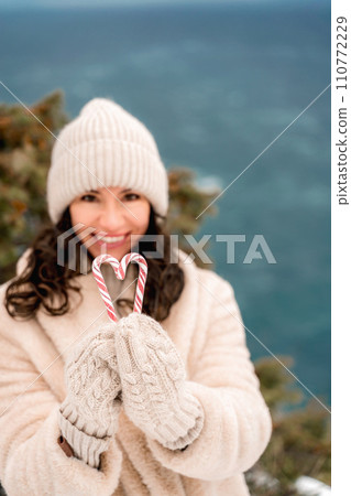 Woman candy sea. Smiling woman in knitted hat, mittens and beige coat holding lollipops candy canes in her hands in shape of heart against the backdrop of the sea. Woman candy sea. Smiling woman in knitted hat, mittens and beige coat holding lollipops candy canes in her hands in shape of heart against the backdrop of the sea. 110772229