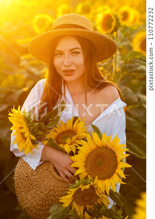A girl in a hat on a beautiful field of sunflowers against the sky in the evening light of a summer sunset. Sunbeams through the flower field. Natural background. A girl in a hat on a beautiful field of sunflowers against the sky in the evening light of a summer sunset. Sunbeams through the flower field. Natural background. 110772232