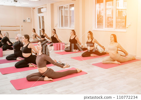 A group of six athletic women doing pilates or yoga on pink mats in front of a window in a beige loft studio interior. Teamwork, good mood and healthy lifestyle concept. 110772234