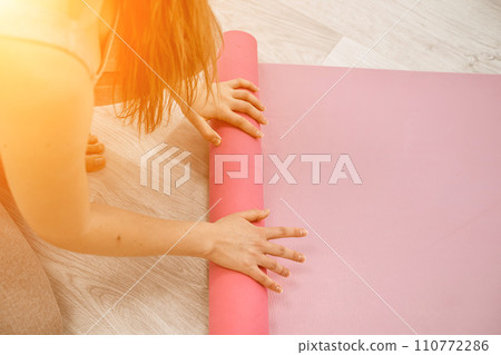 A young woman rolls a pink fitness or yoga mat before or after exercising, exercising at home in the living room or in a yoga studio. Healthy habits, keep fit, weight loss concept. Closeup photo 110772286