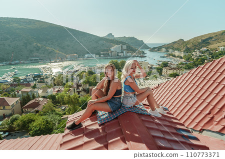 Two women sitting on a red roof, enjoying the view of the town and the sea. Rooftop vantage point. In the background, there are several boats visible on the water, adding to the picturesque scene. Two women sitting on a red roof, enjoying the view of the town and the sea. Rooftop vantage point. In the background, there are several boats visible on the water, adding to the picturesque scene. 110773371