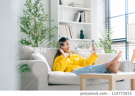 Happy young asian woman wearing headset and relaxing on comfortable couch at home Happy young asian woman wearing headset and relaxing on comfortable couch at home 110773655