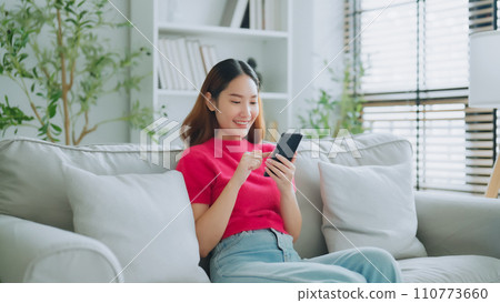 Happy young asian woman relax on comfortable couch at home texting messaging on smartphone Happy young asian woman relax on comfortable couch at home texting messaging on smartphone 110773660