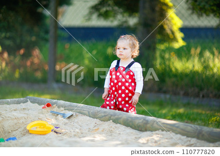 Cute toddler girl playing in sand on outdoor playground. Beautiful baby in red gum trousers having fun on sunny warm summer day. Child with colorful sand toys. Healthy active baby outdoors plays games Cute toddler girl playing in sand on outdoor playground. Beautiful baby in red gum trousers having fun on sunny warm summer day. Child with colorful sand toys. Healthy active baby outdoors plays games 110777820