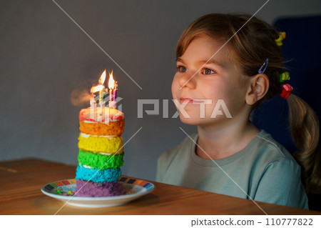 Happy little preschool girl celebrating birthday. Closeup of child with homemade rainbow cake, indoor. Happy healthy toddler blowing six candles on cake. Selective focus on cake Happy little preschool girl celebrating birthday. Closeup of child with homemade rainbow cake, indoor. Happy healthy toddler blowing six candles on cake. Selective focus on cake 110777822