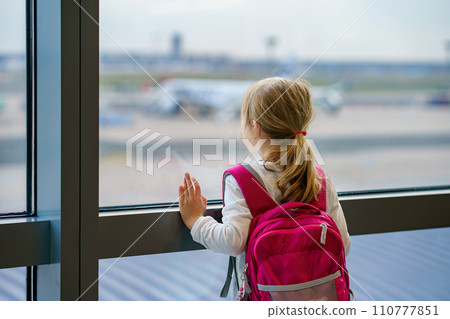 Little Girl at the Airport Waiting for Boarding at the Big Window. Cute Kid Stands at the Window against the Backdrop of Airplanes. Looking Forward to Leaving for a Family Summer Vacation 110777851