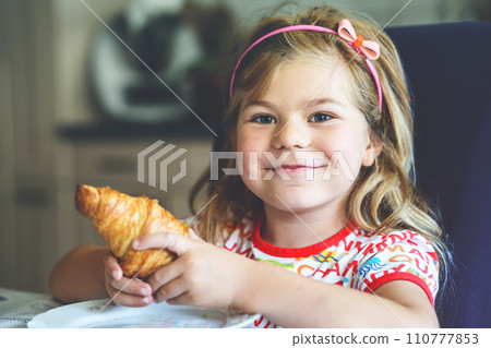 Smiling child at breakfast. Food and happy kids. The girl is eating a croissant. Cute preschool girl having healthy meal. 110777853
