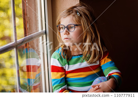 Adorable little preschool girl with eyeglasses sitting by the window. Thoughtful child looking out. Lonely kid. Adorable little preschool girl with eyeglasses sitting by the window. Thoughtful child looking out. Lonely kid. 110777911