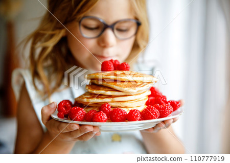 Little happy preschool girl with a large stack of pancakes and raspberries for breakfast. Positive child eating healthy homemade food in the morning. Little happy preschool girl with a large stack of pancakes and raspberries for breakfast. Positive child eating healthy homemade food in the morning. 110777919