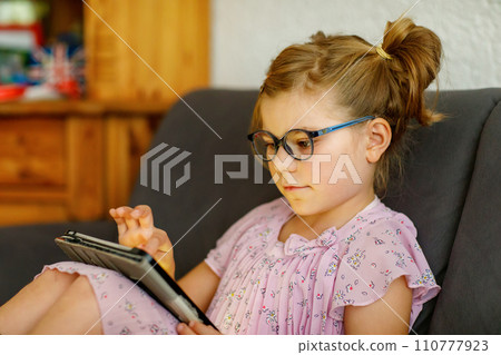 Positive little girl, holding tablet computer in her hands. Preschool child with eyeglasses playing games and learning with different apps. Positive little girl, holding tablet computer in her hands. Preschool child with eyeglasses playing games and learning with different apps. 110777923