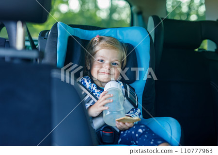 Adorable baby girl with blue eyes sitting in car safety seat. Toddler child going on family vacations and jorney. Smiling happy child during traffic jam, drinking milk from bottle and eating bisquit 110777963