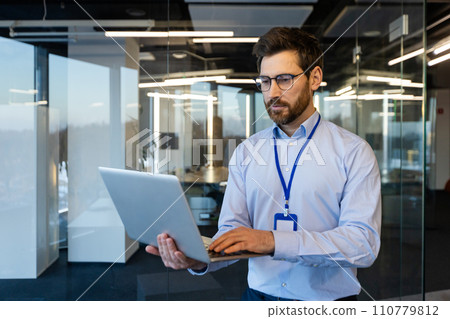 Serious businessman, system administrator stands with a laptop in his hands, a thinking man in a shirt reads data thoughtfully, near the window, a programmer works inside the office with a computer. 110779812