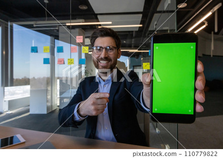 Businessman inside office showing green phone screen recommend app on phone, man in business suit, boss investor working on laptop. Businessman inside office showing green phone screen recommend app on phone, man in business suit, boss investor working on laptop. 110779822