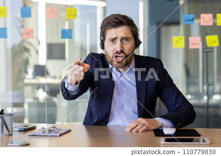 Webcam view, angry man with headset phone yelling at webcam, computer screen, businessman arguing with colleagues and partners inside office at workplace, video call online meeting with employees. 110779830