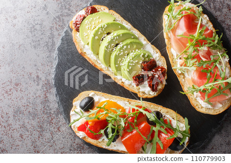 Three different open sandwiches with arugula, jamon, sun-dried tomatoes, avocado, cream cheese, olives and roasted peppers close-up on a slate board. Horizontal top view 110779903
