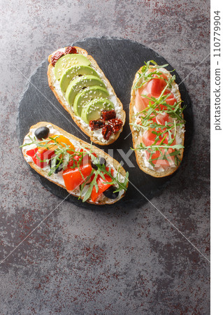 Fresh open sandwiches with arugula, jamon, sun-dried tomatoes, avocado, cream cheese, olives and roasted peppers close-up on a slate board. Vertical top view Fresh open sandwiches with arugula, jamon, sun-dried tomatoes, avocado, cream cheese, olives and roasted peppers close-up on a slate board. Vertical top view 110779904