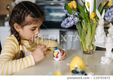 Cute four year girl in beige apron sitting on kitchen table and painting easter eggs 110783872