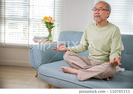 Lifestyle senior man sitting on sofa in living room holding hands in mudra practicing home yoga in lotus pose, Elderly relaxed man do physical exercises following healthy on stress free weekend 110785153