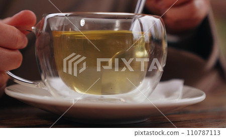 Lady stirs green tea with spoon in transparent glass cup on wooden table in park cafe. Lady takes tea break rotating spoon to mix sugar. Tea for maintaining lady figure stirring hot drink with spoon 110787113