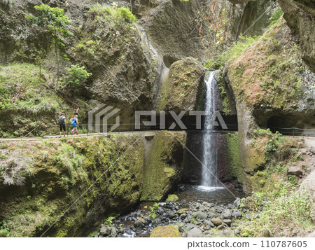 View of waterfall crossing the path at levada, water irrigation channel and tropical plants at hiking trail Levada do moinho to levada nova waterfall. Madeira, Portugal 110787605