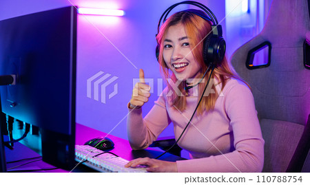 Gamer playing online game wear gaming headphones looking to camera expressing success with game giving thumbs up sign, Smiling woman live stream she play video game at home neon lights living room Gamer playing online game wear gaming headphones looking to camera expressing success with game giving thumbs up sign, Smiling woman live stream she play video game at home neon lights living room 110788754