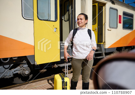 Young woman with backpack and suitcase, standing at railway station, waiting for train, dreamily looking into distance 110788907