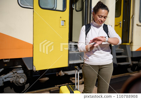 Backpacked female tourist checking time n her wrist watch, standing on the railway station platform against colorful train background 110788908