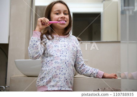 Little kid girl brushing her teeth, standing in the gray minimalist home bathroom, smiling looking at camera. Oral care Little kid girl brushing her teeth, standing in the gray minimalist home bathroom, smiling looking at camera. Oral care 110788935