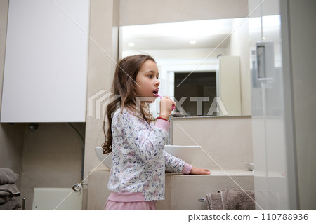 Little kid girl brushing her teeth, standing in the gray minimalist home bathroom. Dental care. Oral hygiene. Copy space 110788936