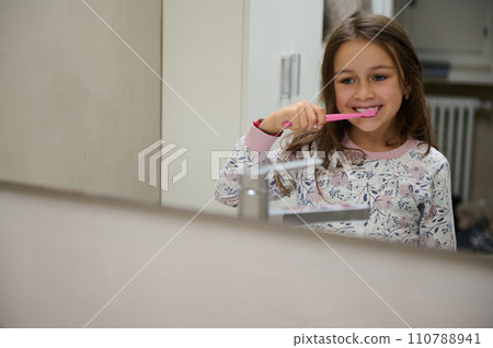 Adorable little kid girl looking at her mirror reflection while brushing teeth in the the bathroom. Healthy lifestyle. Healthy habits. Dental care and hygiene concept 110788941
