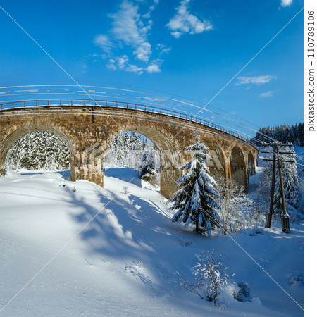 Stone viaduct (arch bridge) on railway through mountain snowy fir forest. Snow drifts on wayside and hoarfrost on trees and electric line wires. Stone viaduct (arch bridge) on railway through mountain snowy fir forest. Snow drifts on wayside and hoarfrost on trees and electric line wires. 110789106